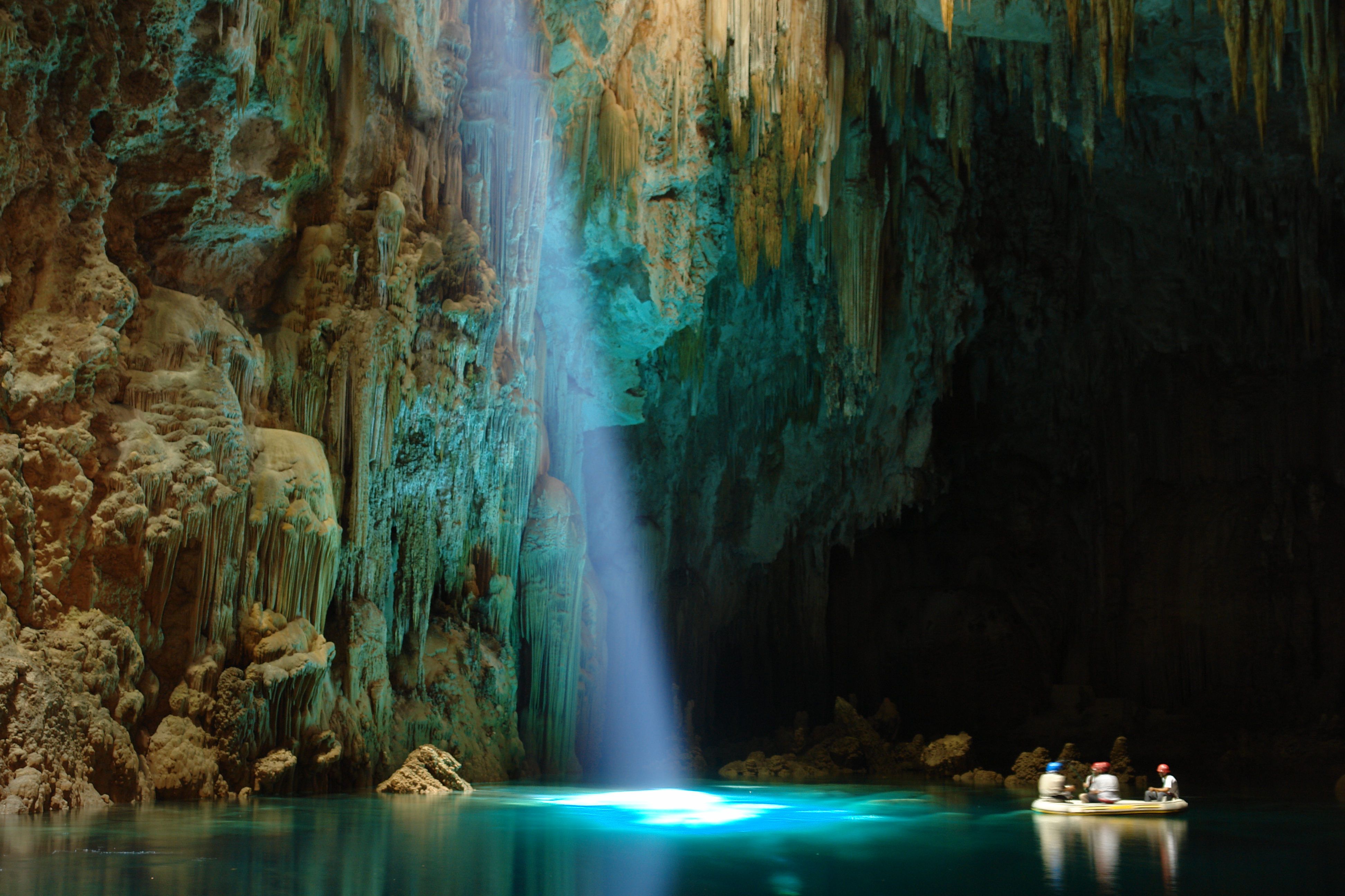 Feixe de luz solar iluminando o lago subterrâneo do Abismo Anhumas em Bonito MS, com turistas em um bote inflável observando as formações rochosas gigantes.