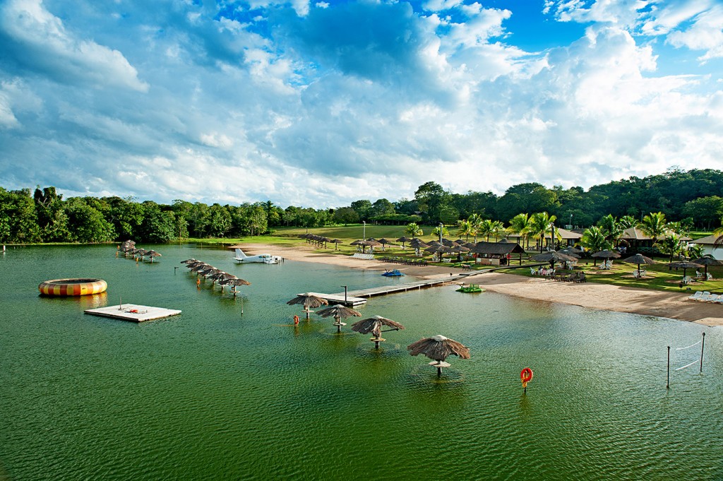 Vista panorâmica do Balneário Praia da Figueira em Bonito MS, mostrando a lagoa de águas verdes, quiosques na areia e um hidroavião estacionado.
