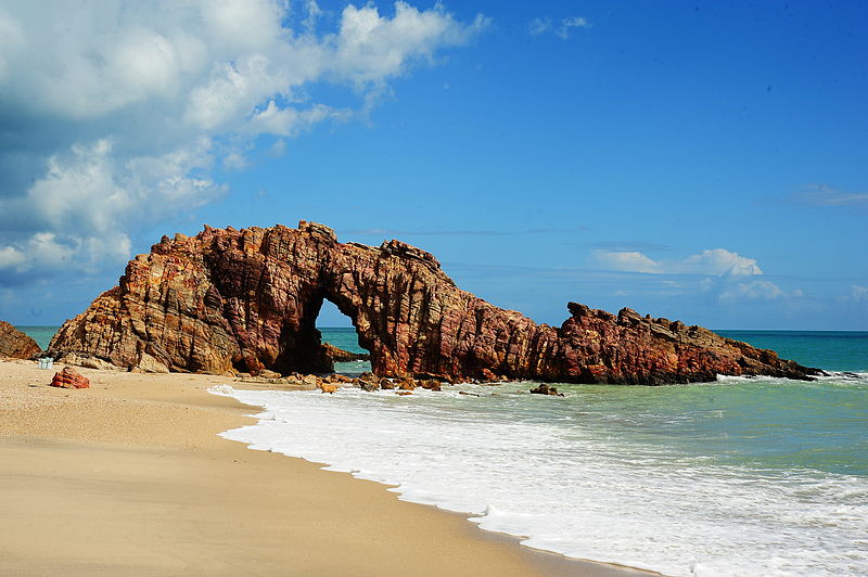 Famosa formação rochosa da Pedra Furada na praia de Jericoacoara, Ceará, com mar azul e céu ensolarado.