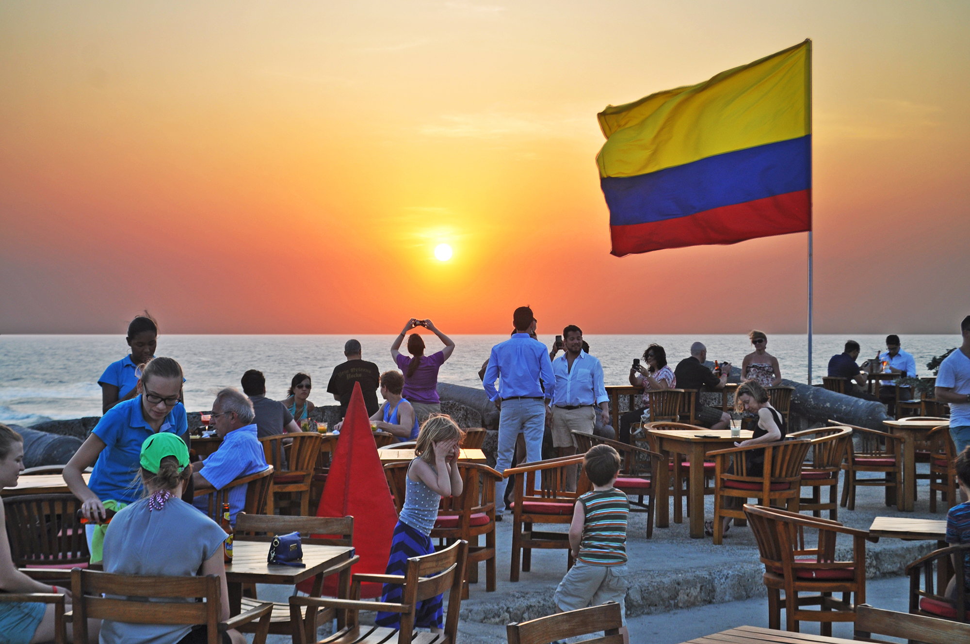 Pôr do sol dourado no famoso bar Café del Mar, situado sobre as muralhas de Cartagena, com a bandeira da Colômbia tremulando ao vento e turistas observando a vista.