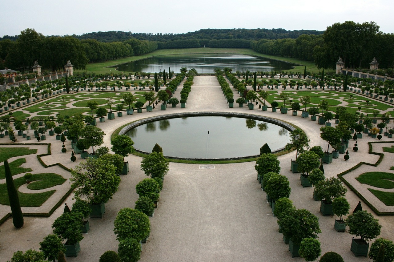 Vista ampla e simétrica dos Jardins do Palácio de Versalhes, mostrando os caminhos geométricos, arbustos podados e o lago ao fundo.