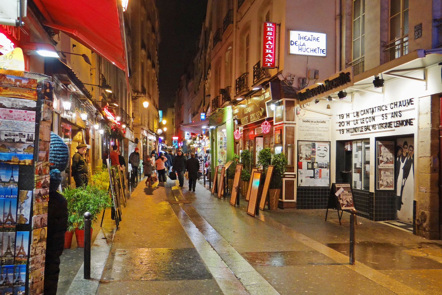 A movimentada Rue de la Huchette à noite no Quartier Latin, repleta de restaurantes iluminados, letreiros de neon e turistas caminhando.