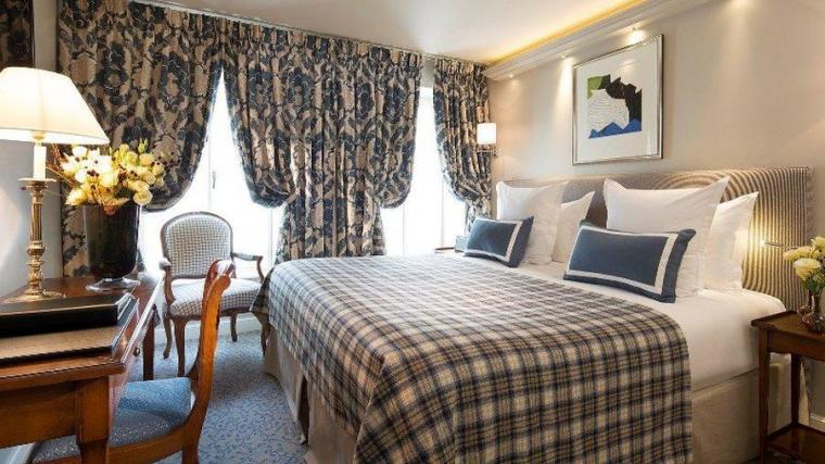 Quarto de hotel elegante em Paris com decoração clássica, roupa de cama xadrez, cortinas pesadas e escrivaninha de madeira antiga.