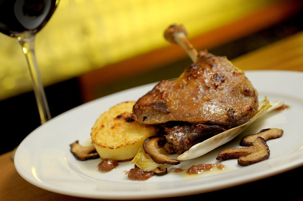 Prato clássico da culinária francesa: coxa de pato confitada (Confit de Canard) servida com batatas e cogumelos, com uma taça de vinho ao fundo.