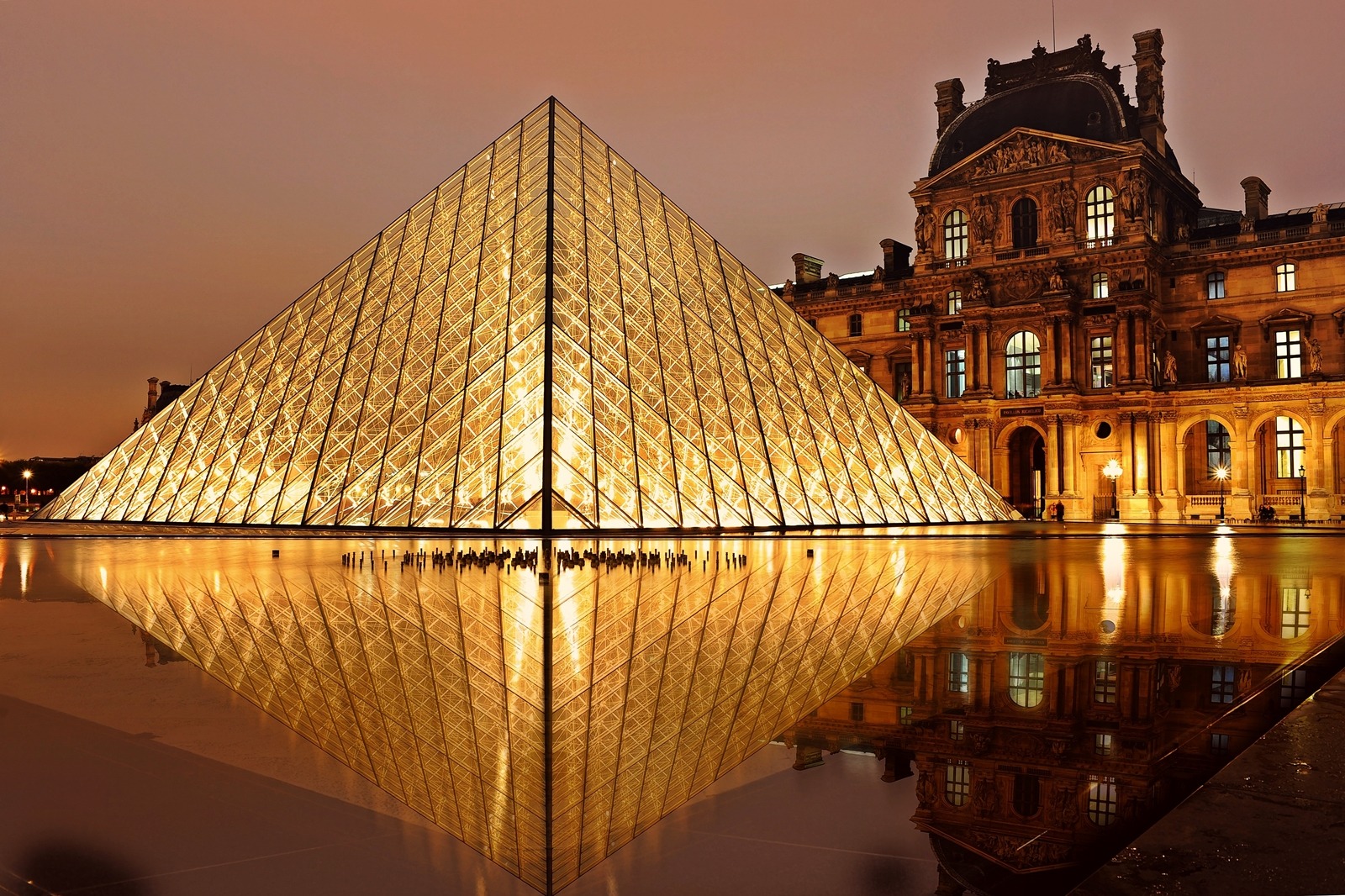 Pirâmide de vidro do Museu do Louvre iluminada à noite, com seu reflexo na água e a fachada clássica do palácio ao fundo.