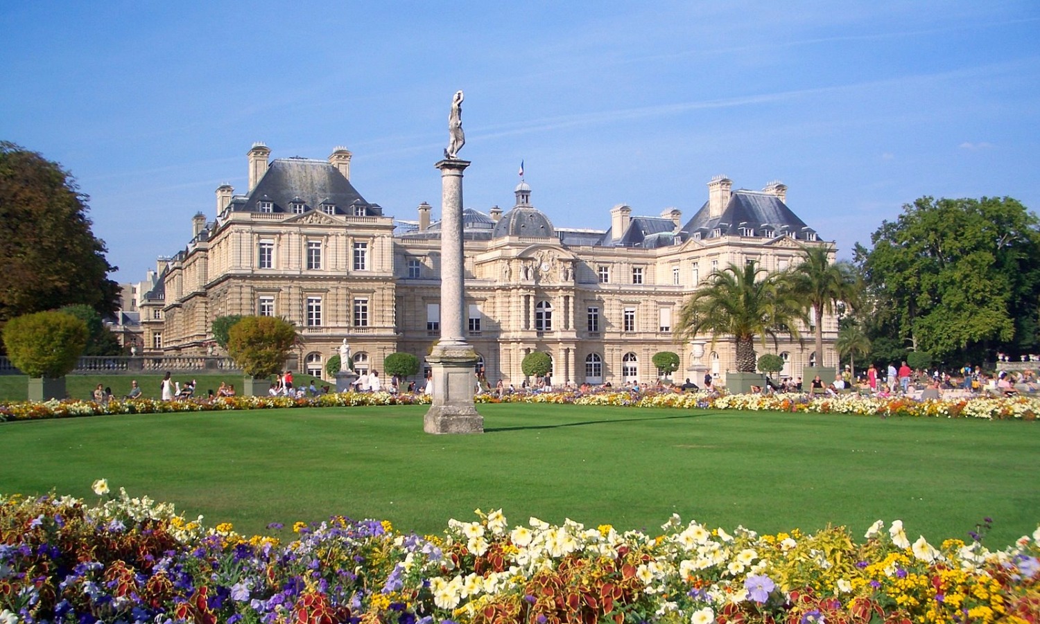 Palácio e Jardins de Luxemburgo em Paris durante o verão, com gramados verdes, canteiros de flores coloridos e estátuas sob o céu azul.