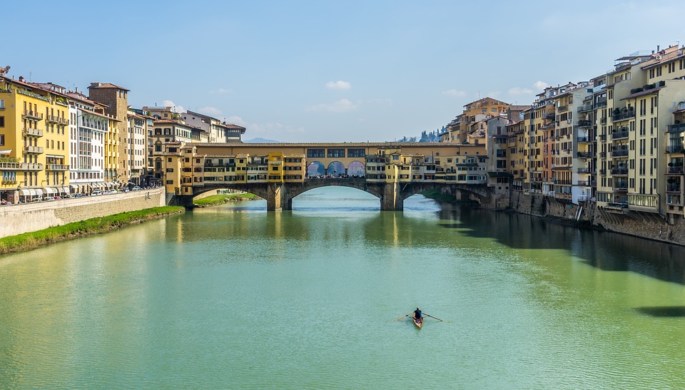 Vista panorâmica da Ponte Vecchio sobre o Rio Arno em Florença, com arquitetura medieval e um pequeno barco navegando.