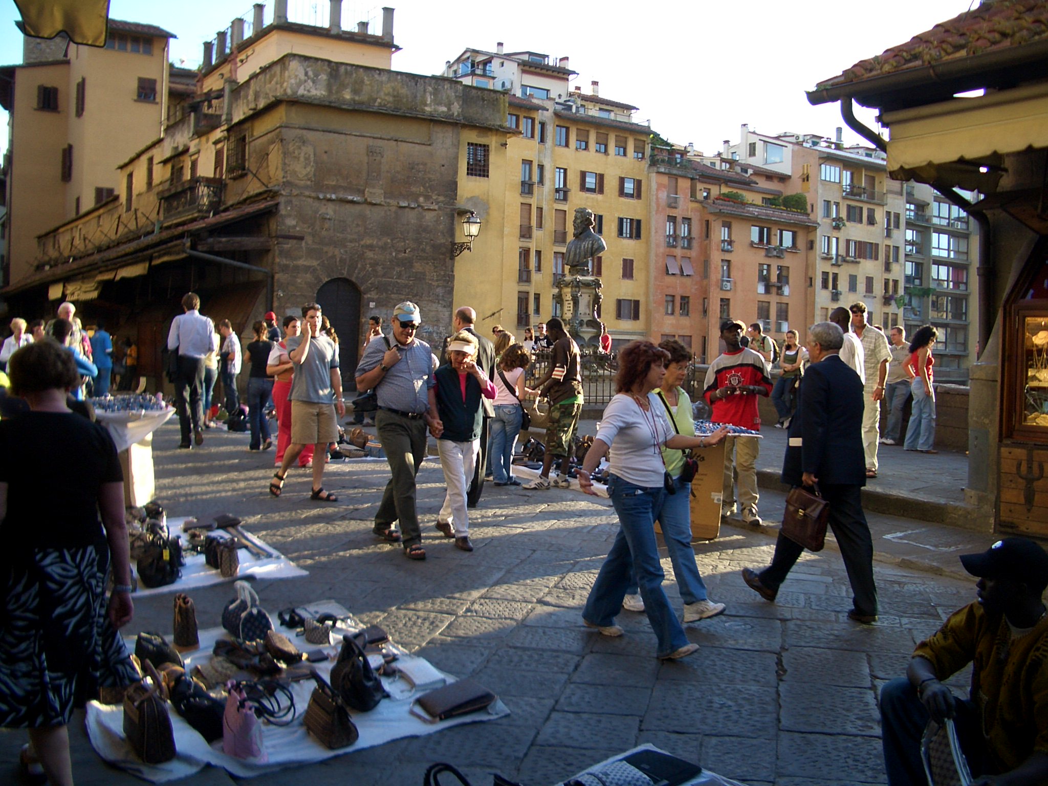 Turistas e vendedores ambulantes caminhando perto da Ponte Vecchio em um dia ensolarado em Florença.