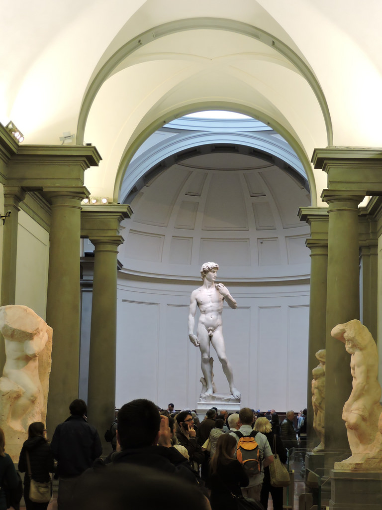 Estátua de David de Michelangelo exposta na Galleria dell'Accademia em Florença, com turistas observando a obra renascentista.
