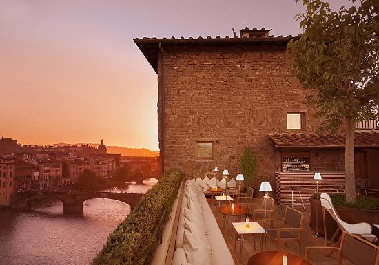 Bar no terraço (rooftop) em Florença com mesas elegantes e vista para o pôr do sol sobre o Rio Arno e a Ponte Vecchio.