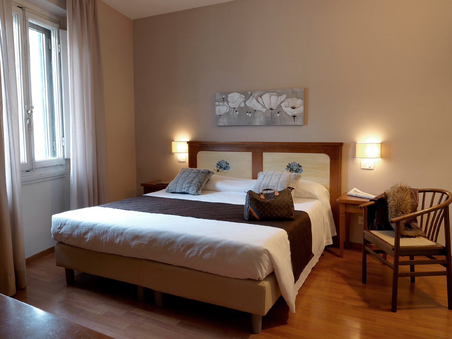 Interior de um quarto de hotel aconchegante e iluminado, com cama de casal grande, piso de madeira e decoração clássica, ideal para descanso durante a viagem.